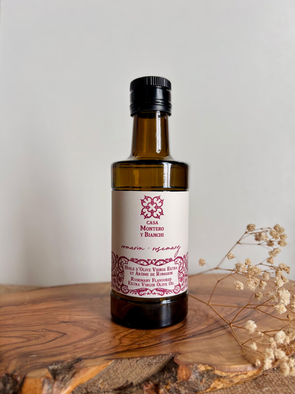 Huile d'olive vierge extra aromatisée au romarin Casa Montero y Bianchi présentée sur une table en bois avec des fleurs séchées.